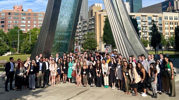 Group of summer associates outdoors at base of art feature with city environment in background